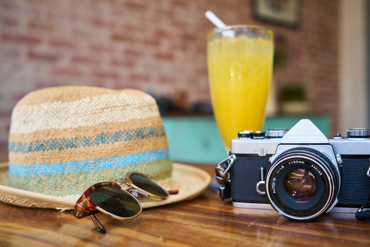 A imagem mostra a borda de uma piscina em segundo plano, e em primeiro plano, um chapéu de praia, óculos de sol, uma taça de suco de laranja e uma máquina fotográfica para registrar os melhores momentos do verão.